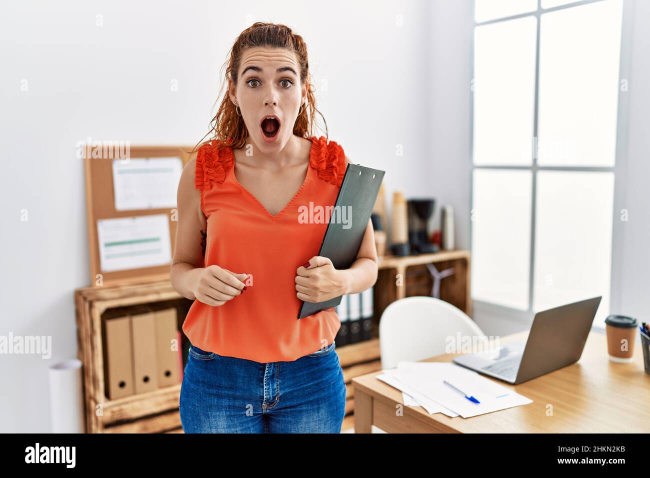 Jeune femme à tête rouge au bureau tenant le presse-papiers effrayée et  stupéfait avec la bouche ouverte pour la surprise, le visage incrédulé  Photo Stock - Alamy