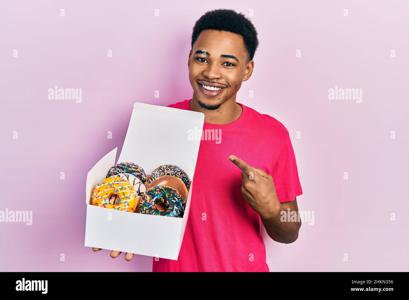 Jeune homme afro-américain tenant une boîte avec de savoureux beignets colorés souriant souriant avec la main et le doigt Banque D'Images