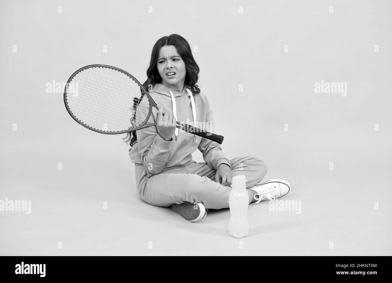 un enfant malheureux s'assoit avec une bouteille d'eau de raquette. un enfant avec une raquette de tennis. une jeune fille va boire de l'eau Banque D'Images