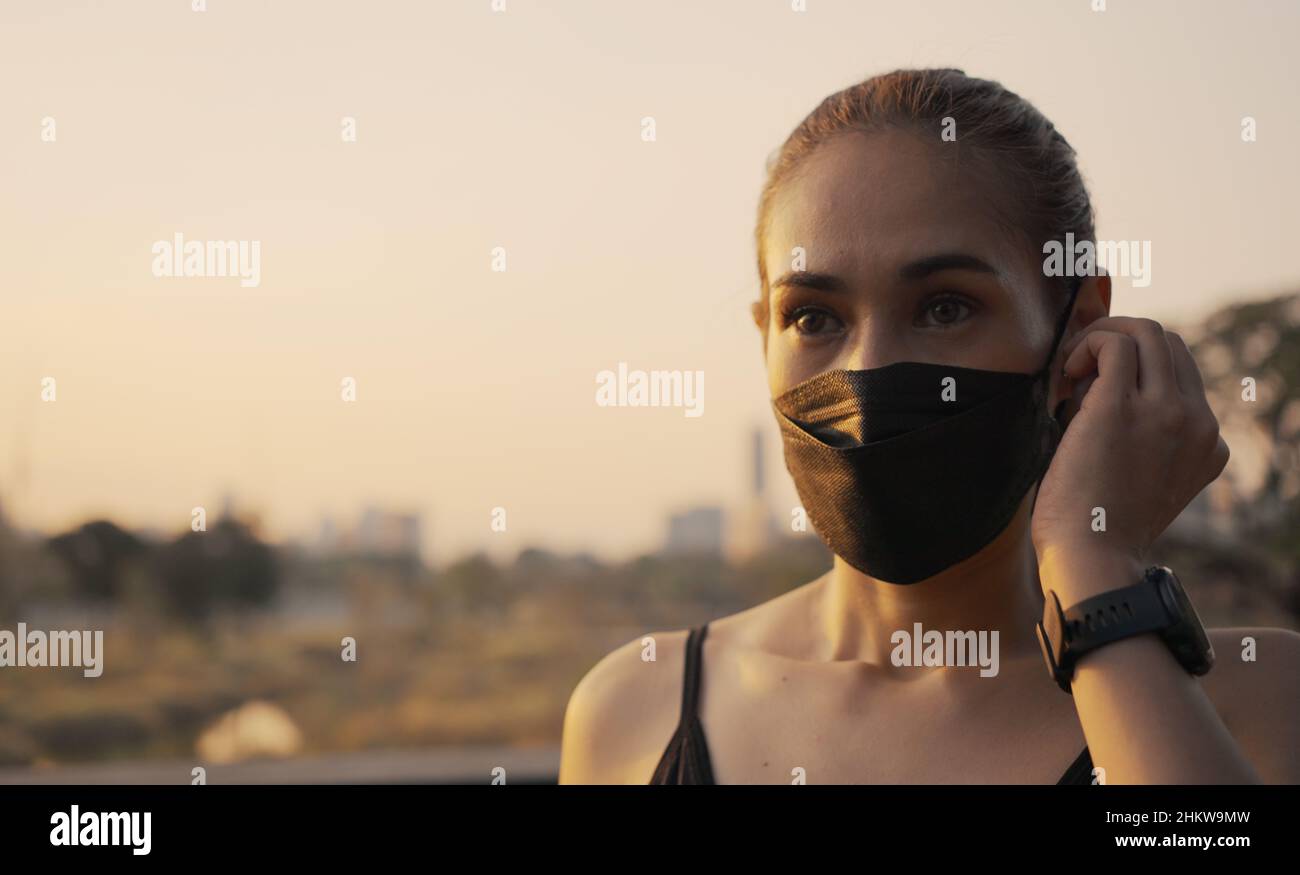 Femme en vêtements de sport portant un masque de protection et des écouteurs sans fil avant de commencer à courir dans la ville au coucher du soleil. Banque D'Images