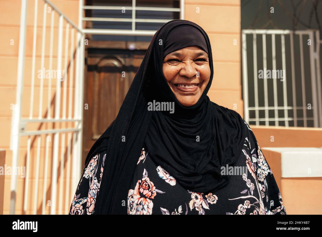 Bonne femme musulmane souriant à la caméra en se tenant debout devant sa maison pendant la journée.Femme d'âge moyen insouciante portant un hijab et Banque D'Images