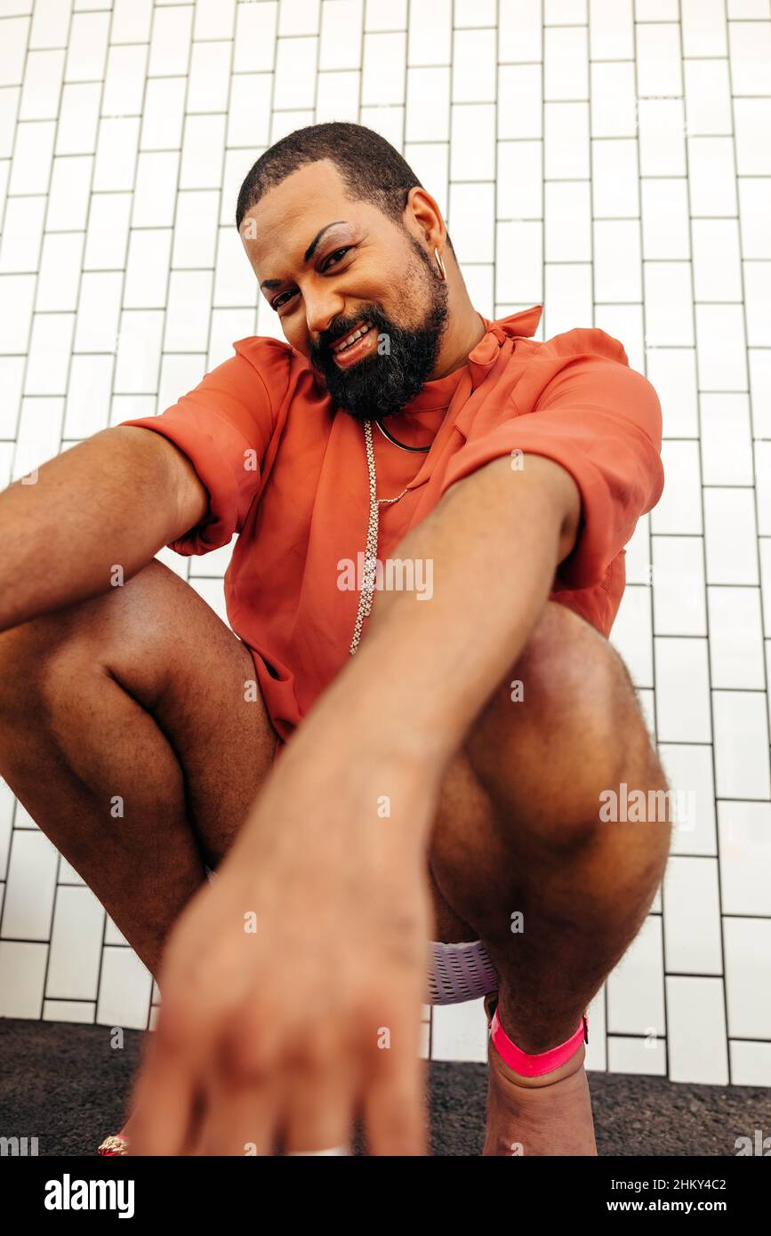 Joyeux jeune, traînez la reine en souriant à l'appareil photo tout en vous squatant sur un fond blanc.Jeune homme non conforme portant du maquillage et des bijoux.Confi Banque D'Images
