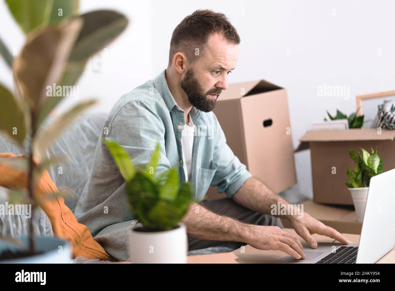 Un homme mûr assis dans une pièce désordonnée avec des boîtes en carton et des plantes, en utilisant un ordinateur portable.Un concept en mouvement. Banque D'Images