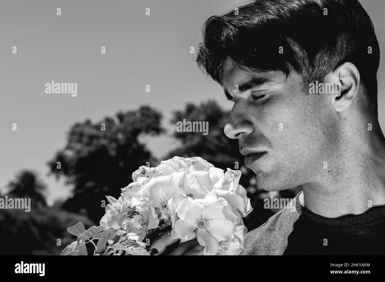 spectaculaire et romantique jeune homme argentin caucasien en noir et blanc avec les yeux fermés odeur d'une fleur dans le parc, avec le ciel sur fond an Banque D'Images