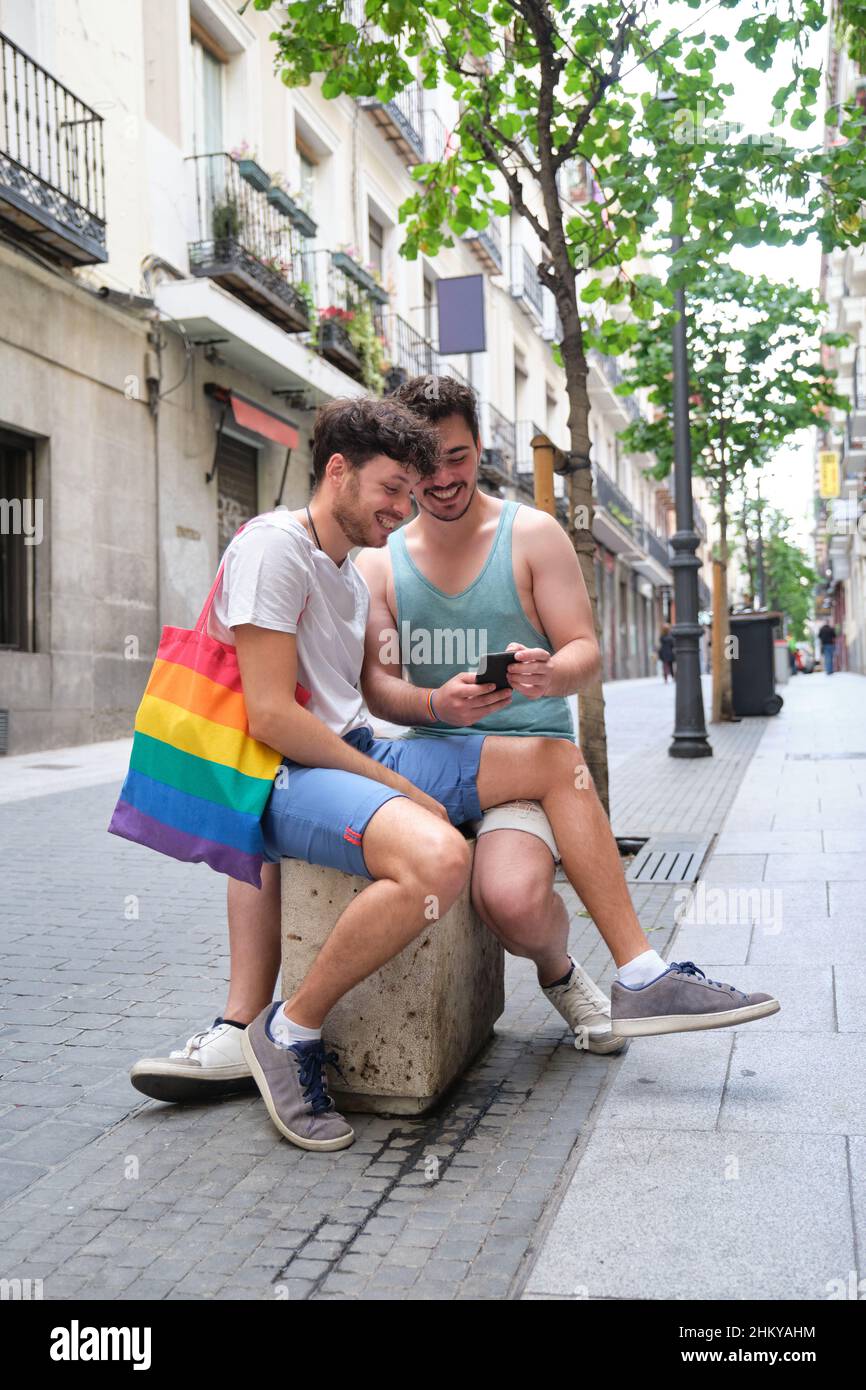 Couple gay utilisant le smartphone et rire assis dans la rue. Banque D'Images