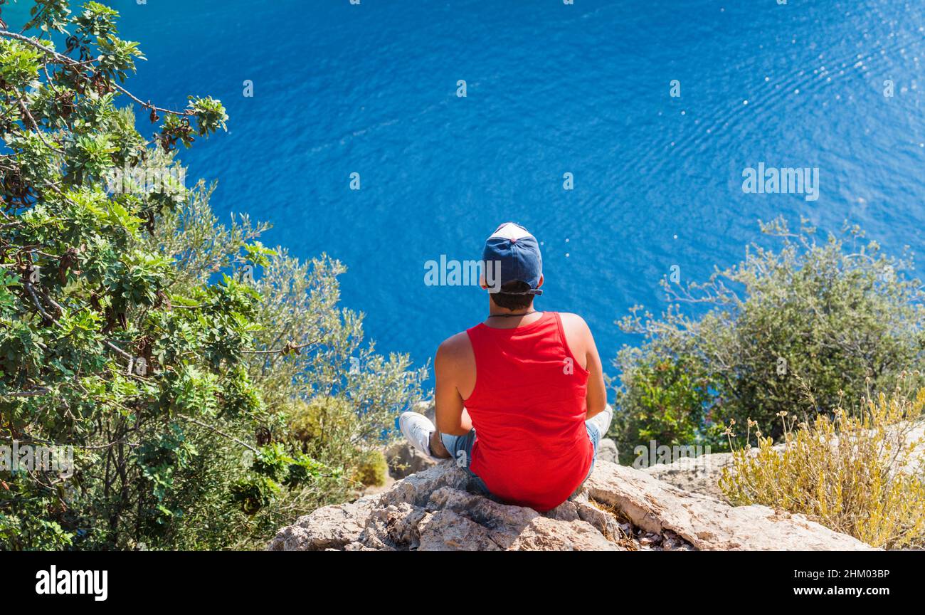 Jeune homme assis au bord d'une falaise et appréciant le magnifique paysage de mer.Vue sur Butterfly Valley en Turquie.Concept de liberté. Banque D'Images