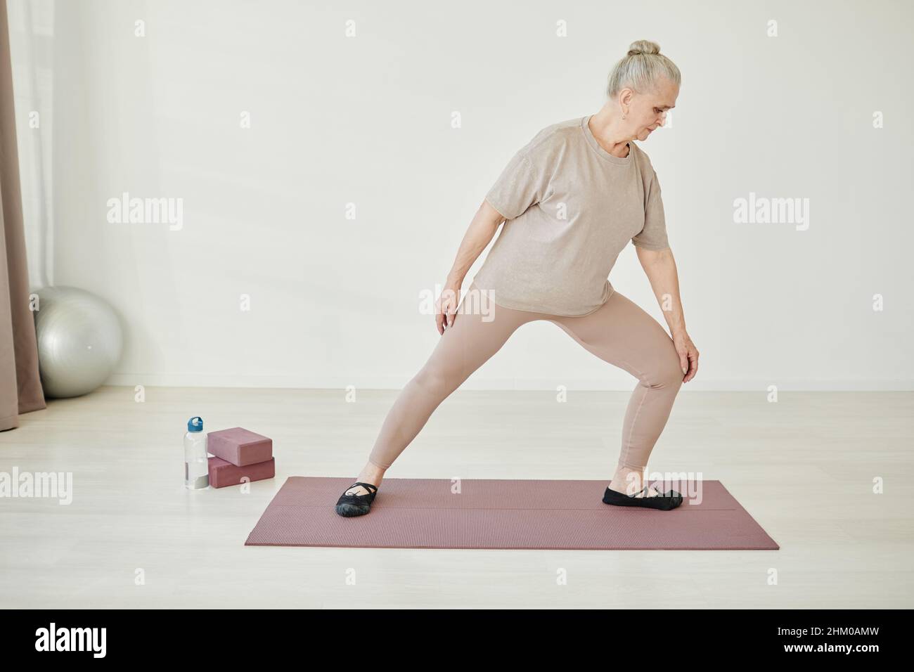 Femme âgée en vêtements d'activité gardant une jambe pliée dans le genou tout en faisant de l'exercice physique sur le tapis dans l'environnement à la maison Banque D'Images