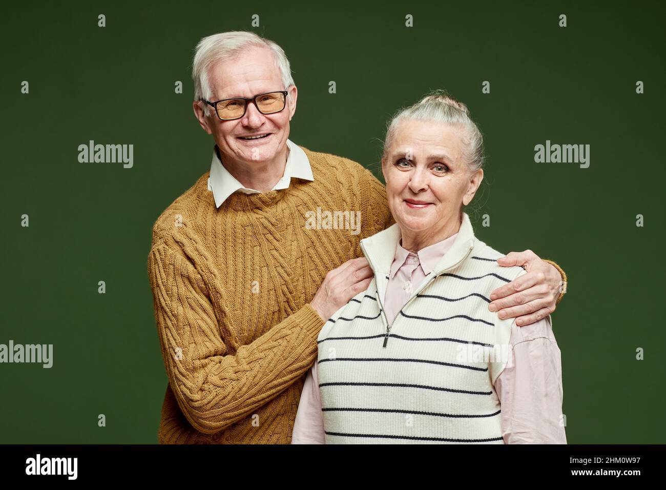 Couple senior contemporain et joyeux, en pull-overs, debout sur fond vert foncé et regardant l'appareil photo avec des sourires Banque D'Images