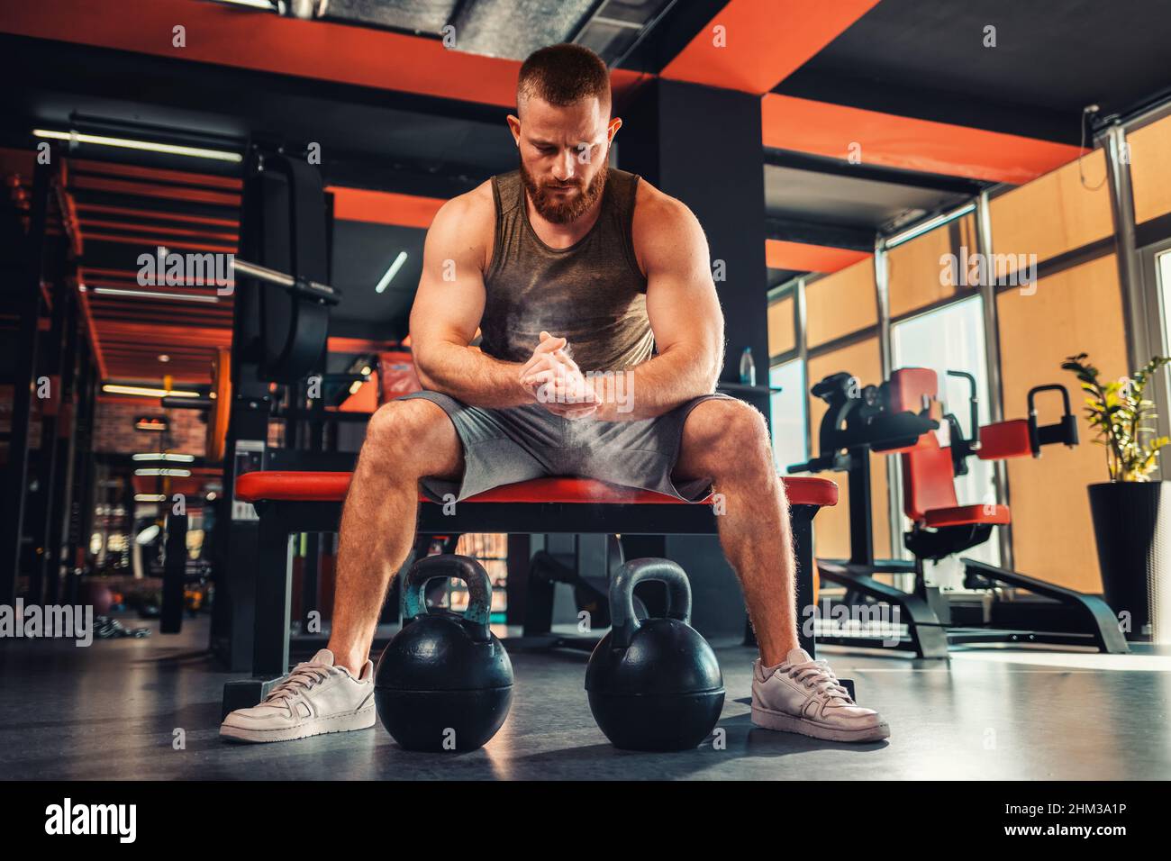 Un jeune homme sportif barbu s'entraîne dans la salle de gym avec des haltères.L'athlète frotte la magnésie dans ses mains.Le concept de forme physique. Banque D'Images