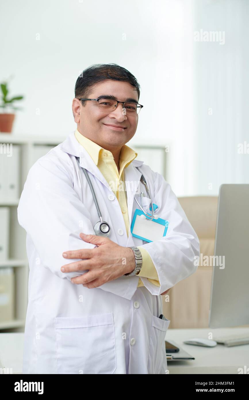Portrait d'un médecin mature positif en blouse de travail blanche croisant les bras et souriant à la caméra Banque D'Images