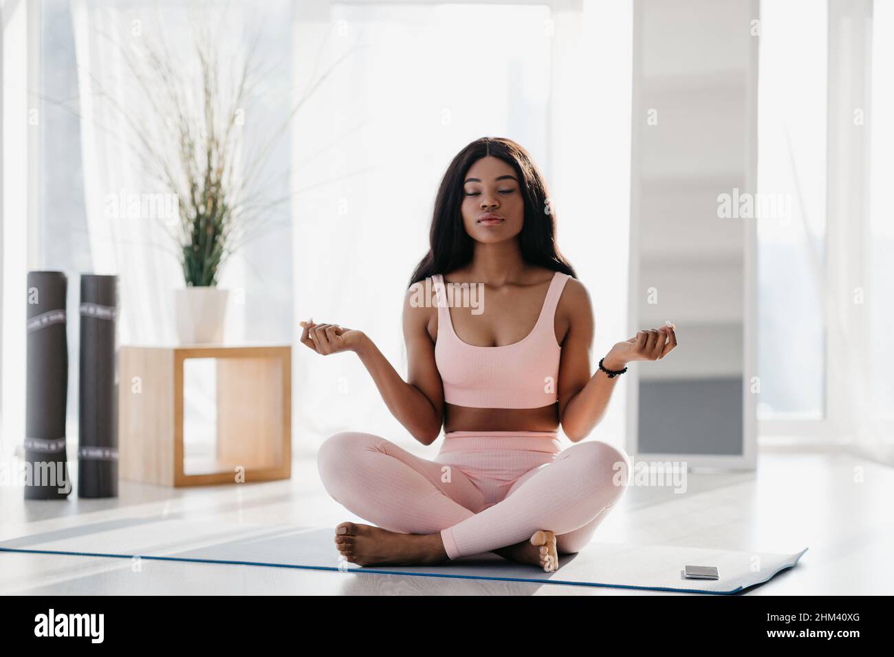 Belle jeune femme noire paisible assise dans la posture du lotus, méditant avec les yeux fermés, pratiquant le yoga sur le tapis à la maison Banque D'Images