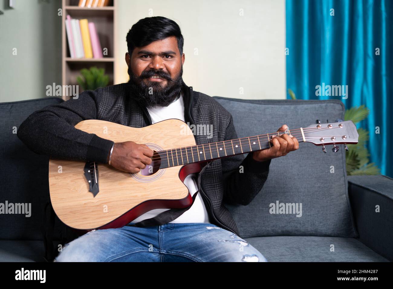 Jeune homme de barbe qui enseigne la guitare en regardant l'appareil photo de la maison - concept de cours de musique en ligne ou virtuelle, passe-temps et artiste professionnel. Banque D'Images