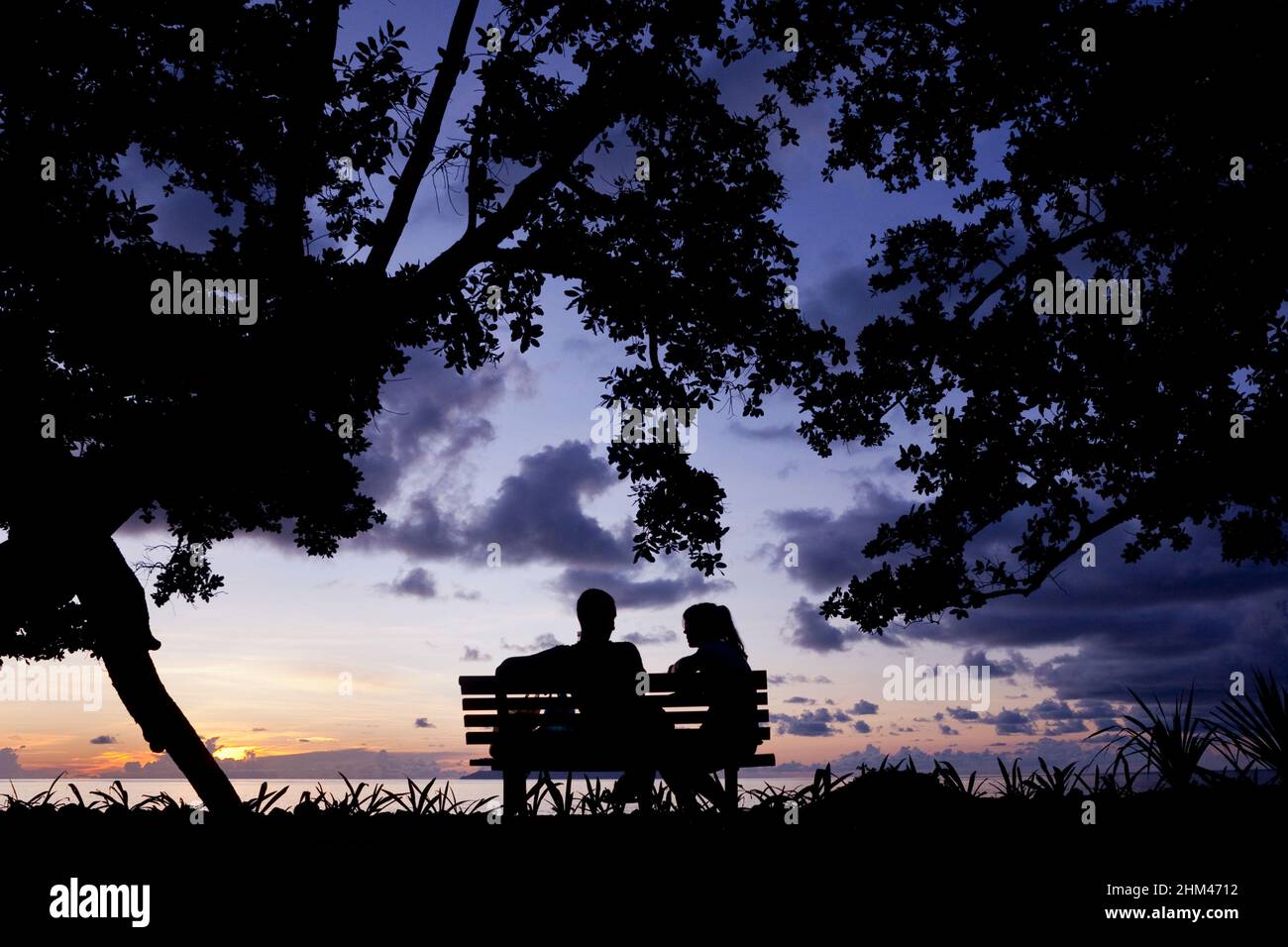 Un couple prenant le coucher du soleil sur la plage de beau Vallon depuis un banc de parc sur l'île de Mahé, Seychelles. Banque D'Images