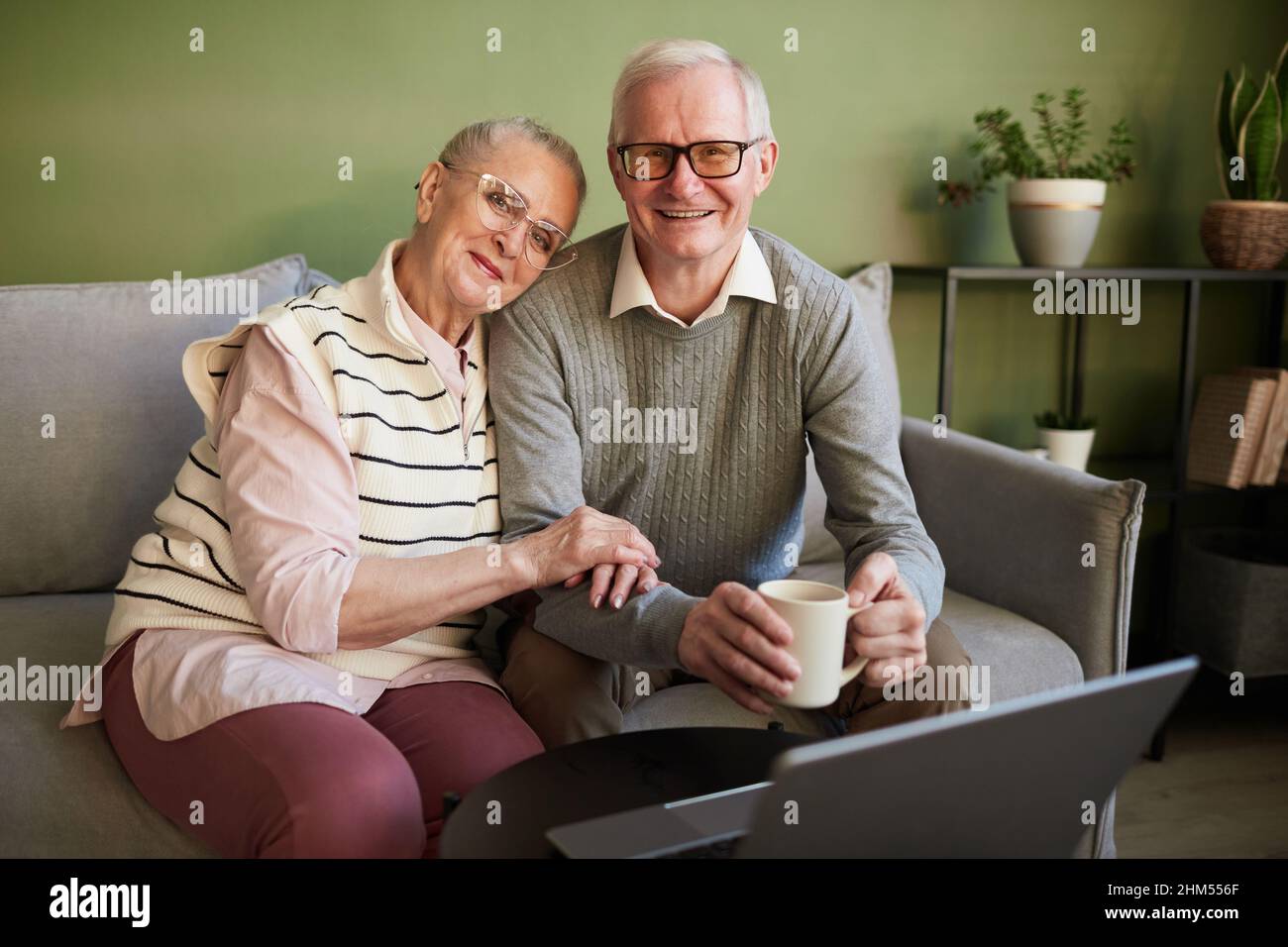 Une femme et un mari senior heureux et affectueux assis sur un canapé devant un ordinateur portable dans le salon et regardant l'appareil photo Banque D'Images