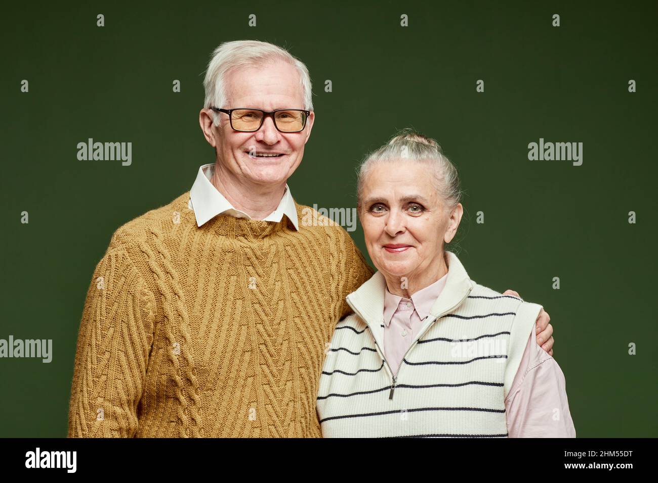 Portrait d'un mari aîné heureux en pull-over marron épousant sa femme en vêtements décontractés à rayures sur fond vert foncé Banque D'Images