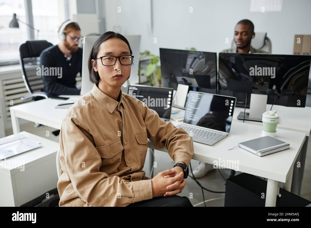 Jeune homme d'affaires asiatique sérieux en lunettes et vêtements décontractés assis près d'un bureau avec deux ordinateurs portables contre ses collègues Banque D'Images