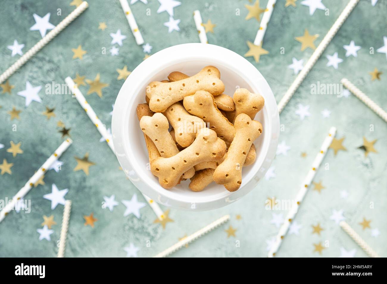 Gâteries pour chiens, biscuits en os dans un bol blanc sur fond vert festif avec étoiles dorées et blanches et pailles pour cocktails. Fête d'anniversaire pour animal de compagnie. Vue de dessus Banque D'Images