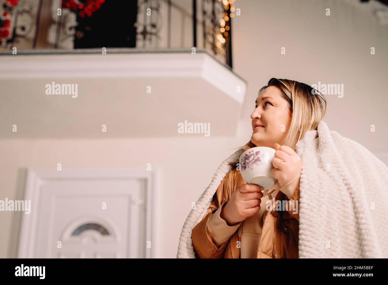 Vue latérale photo d'une femme dehors en vacances d'hiver regardant de côté se tenant près de la maison décorée avec des guirlandes gardant une tasse de boisson chaude. Banque D'Images