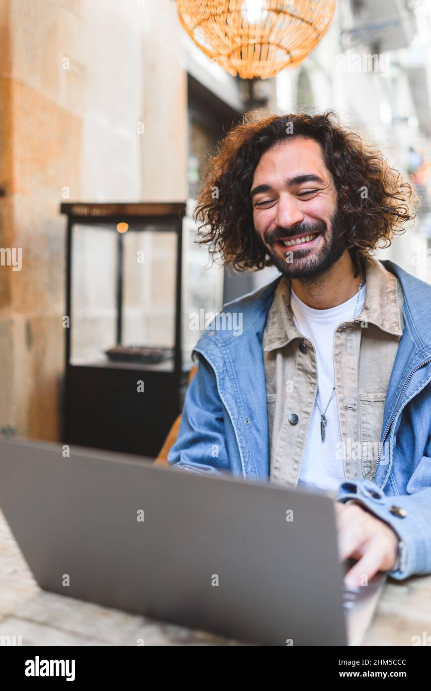 Joyeux hispanique dans des vêtements décontractés avec la barbe et les cheveux bouclés souriant. Il utilise un netbook tout en étant assis à table et en travaillant sur un projet à distance dans un café de rue Banque D'Images