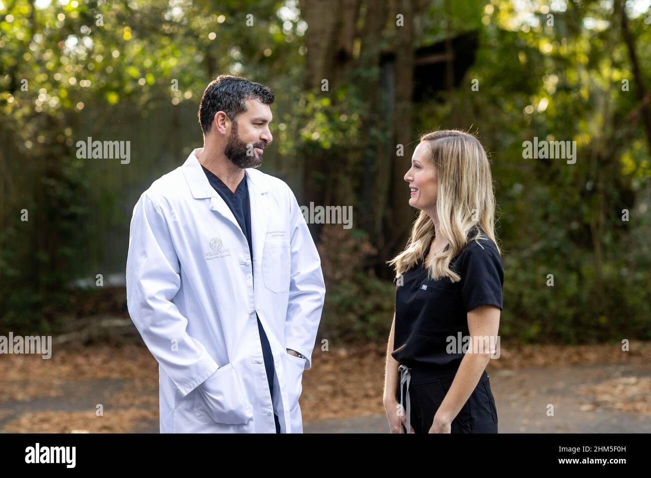 Un médecin aux cheveux foncés et une barbe en gommage noir et un manteau de laboratoire blanc debout à l'extérieur parlant à une infirmière praticienne médicale professionnelle Banque D'Images