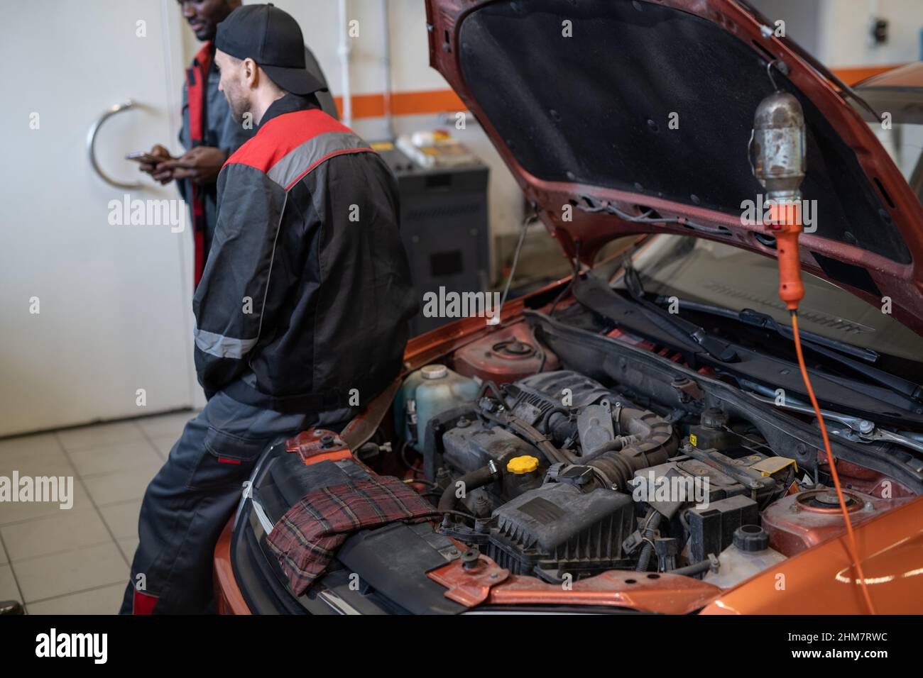 Vue arrière chez un mécanicien de sexe masculin en pause dans un atelier automobile avec capot et moteur ouverts au premier plan, espace de copie Banque D'Images