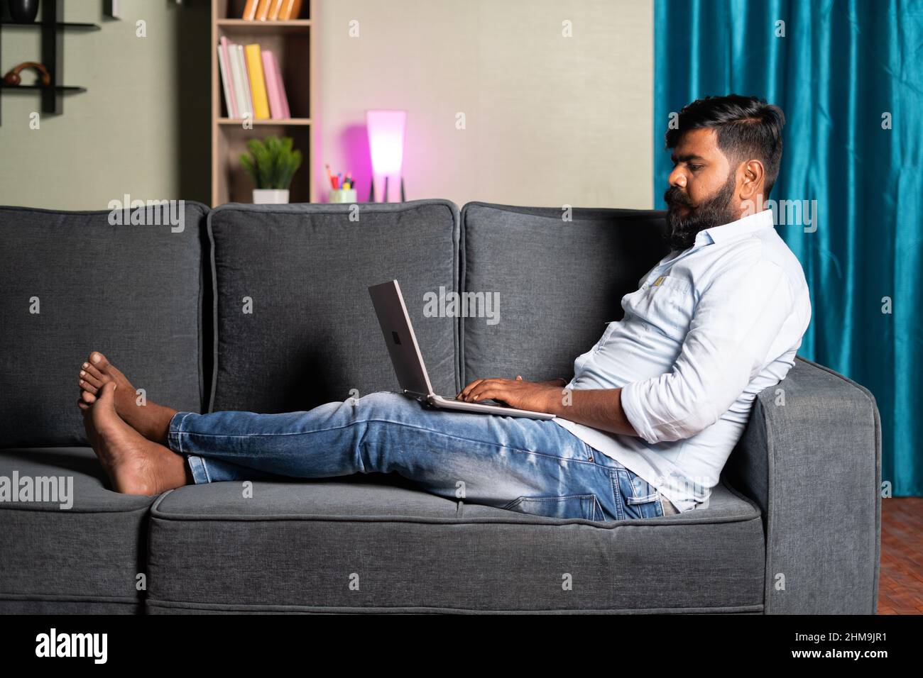 Prise de vue grand angle d'un jeune homme détendu, occupé à utiliser un ordinateur portable assis sur safa à la maison - concept de travail à la maison, nouveau employé normal ou mode de vie professionnel Banque D'Images