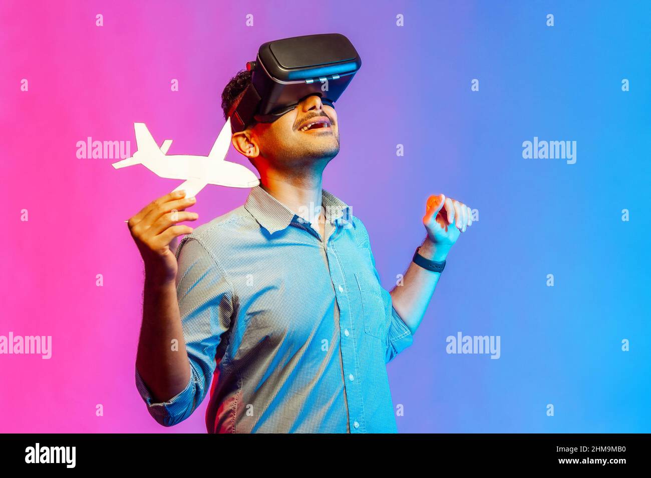 Homme en chemise dans des lunettes VR tenant un maquette d'avion, regardant un vol en réalité augmentée ou des offres de service de compagnie aérienne, des voyages virtuels. Studio d'intérieur isolé sur fond de néon coloré Banque D'Images