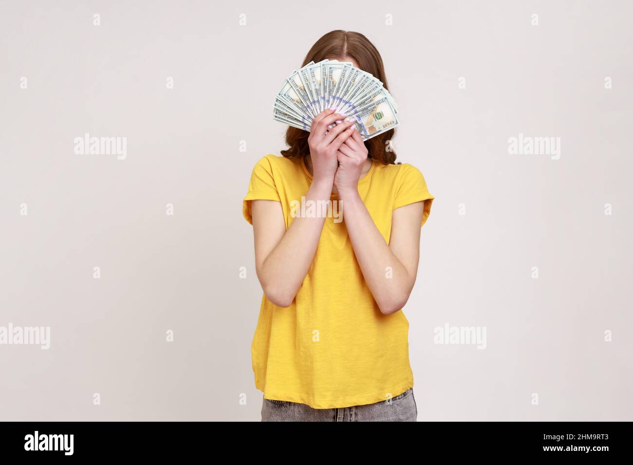 Femme inconnue dans le style urbain jaune T-shirt cache face derrière un bouquet de billets de dollars, personne anonyme tenant de l'argent, gain de loterie, gros profit. Prise de vue en studio isolée sur fond gris. Banque D'Images