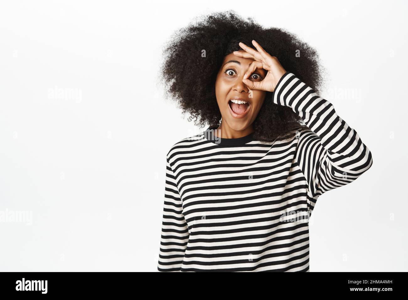 Excitée jeune femme africaine montre OK, zéro geste, ok signe sur l'œil et sourire stupéfait, en regardant smth impressionnant, semble impressionné, blanc Banque D'Images