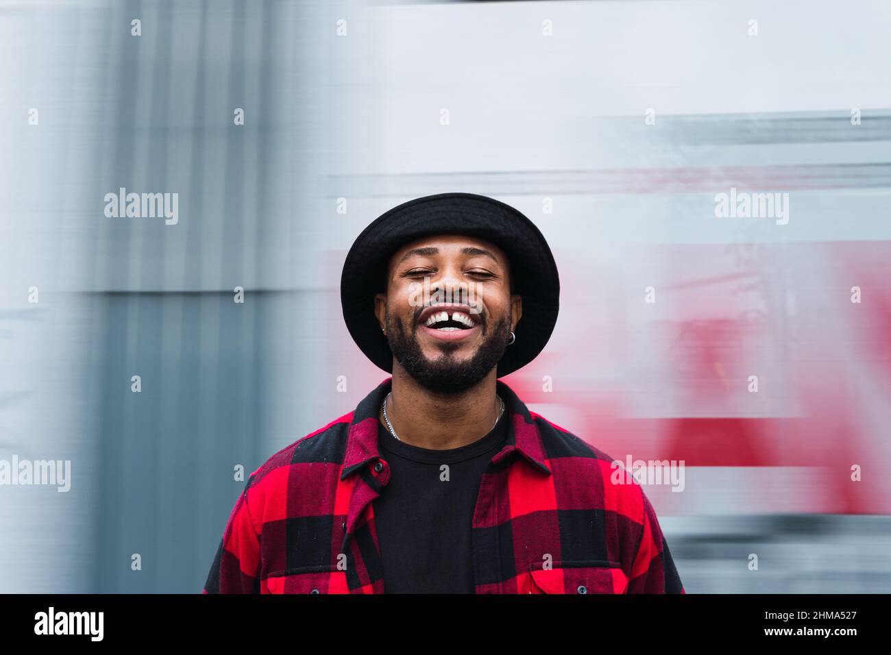 Joyeux homme barbu noir dans une chemise et un chapeau à carreaux regardant l'appareil photo sur un fond flou de la rue de la ville Banque D'Images