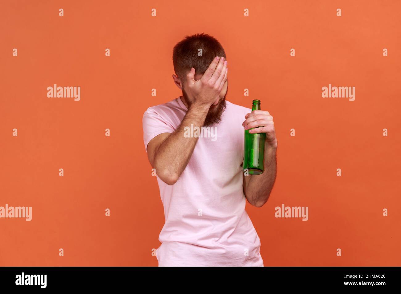 Portrait d'un triste homme barbu debout avec une bouteille de bière, ivre, debout couvrant son visage avec de la paume, portant un T-shirt rose. Studio d'intérieur isolé sur fond orange. Banque D'Images