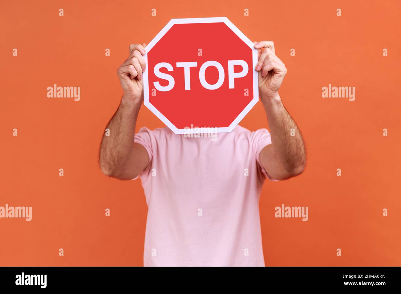 Portrait de l'homme couvrant le visage avec le symbole Stop, anonyme tenant le panneau de signalisation rouge, avertissement à partir, concept d'interdiction, portant un T-shirt rose. Studio d'intérieur isolé sur fond orange. Banque D'Images