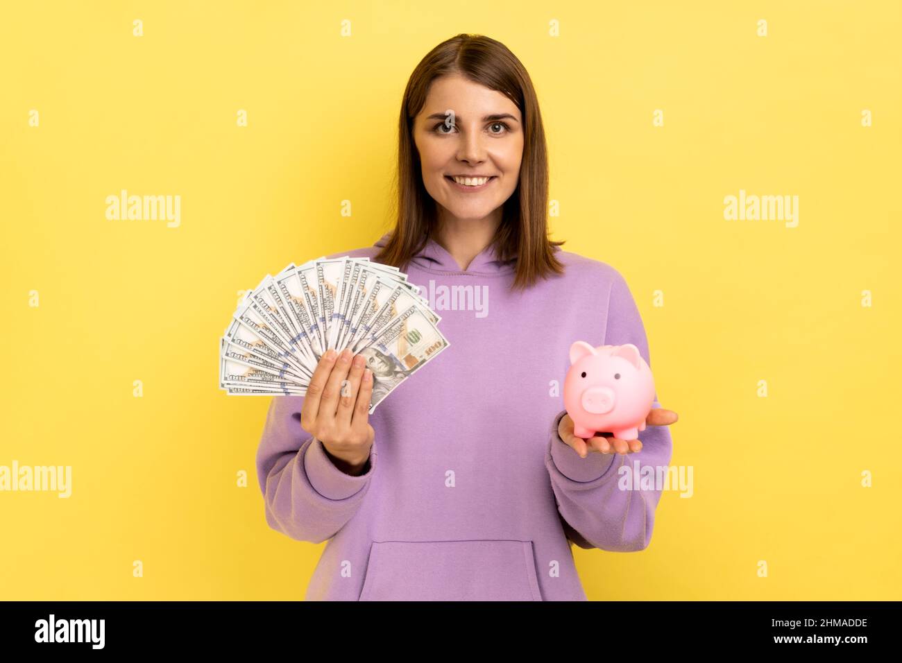 Souriante positive femme avec des cheveux foncés tenant une grande somme d'argent et de la banque de porc, investissement rentable, portant le chandails pourpre. Studio d'intérieur isolé sur fond jaune. Banque D'Images