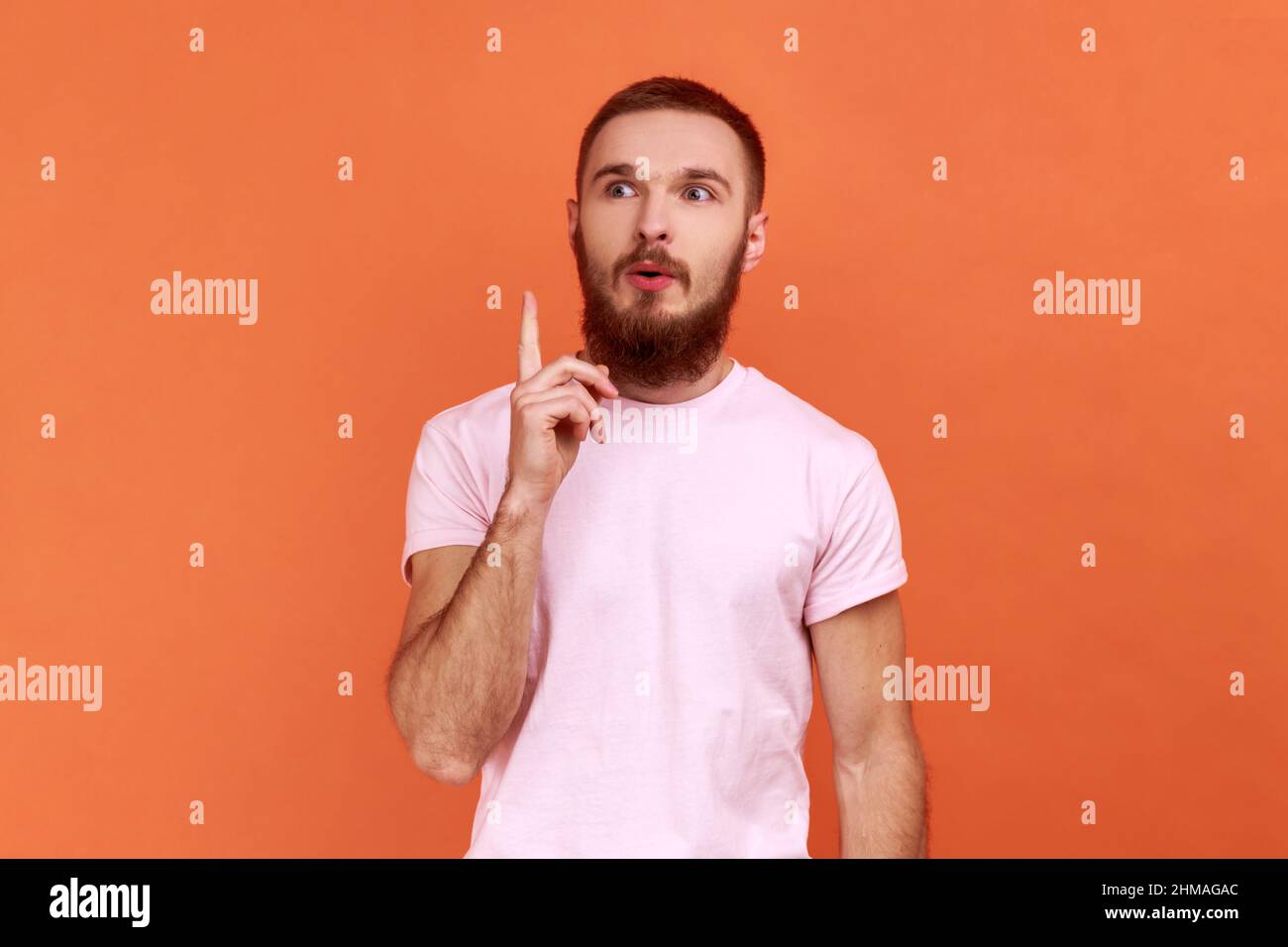 Portrait d'un beau barbu inspiré homme pointant du doigt vers le haut et regardant stupéfait de génie soudain idée, a la solution, portant le T-shirt rose. Studio d'intérieur isolé sur fond orange. Banque D'Images