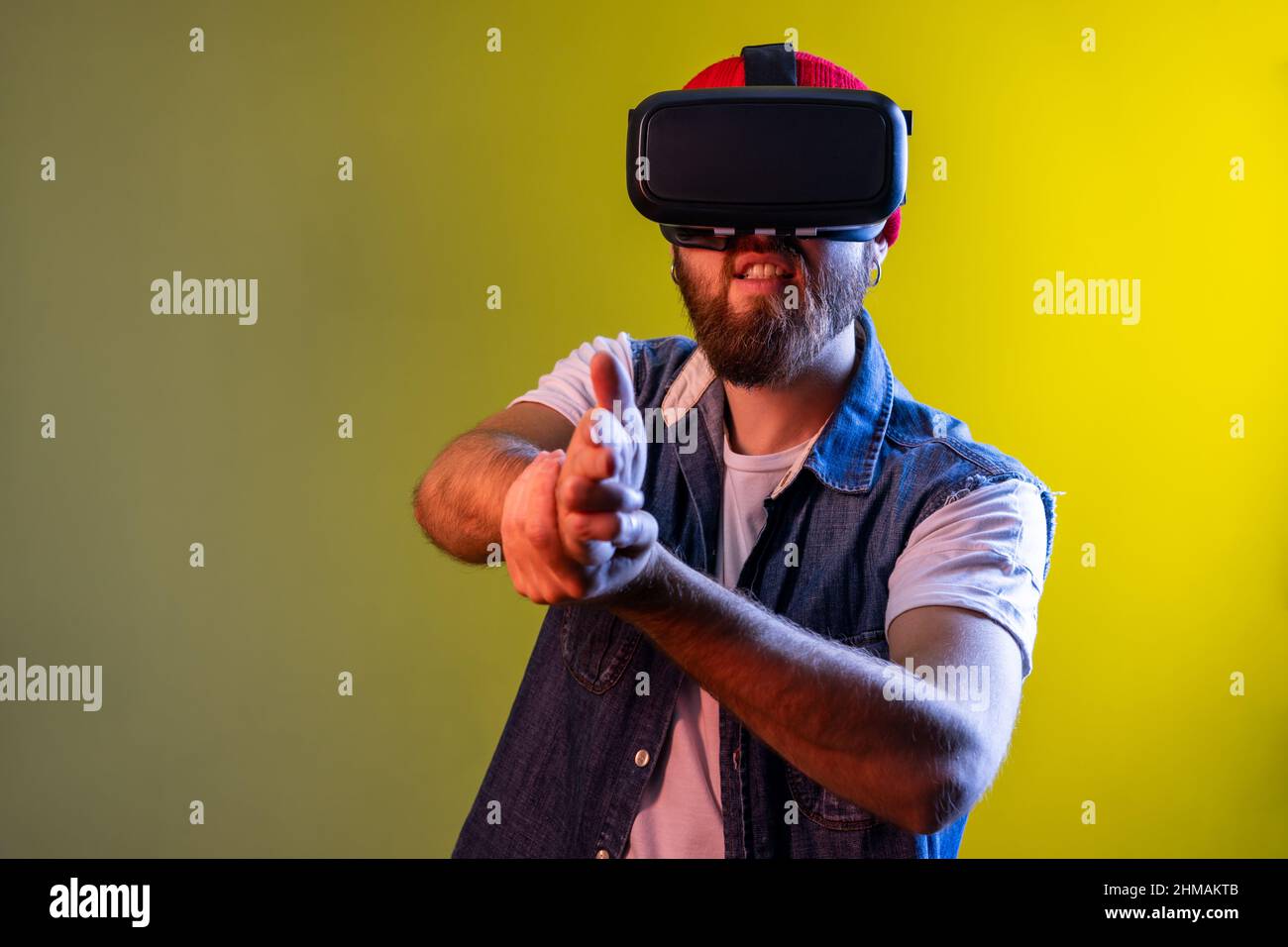 Homme avec des lunettes de réalité virtuelle à la tête jouant au jeu de tir, pointant le doigt du fusil et de tir directement sur la cible, portant une veste en denim. Studio d'intérieur isolé sur fond de néon coloré. Banque D'Images