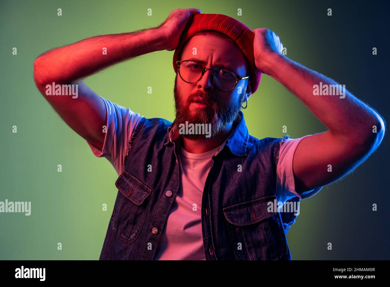 Jeune adulte barbu hipster homme dans des lunettes, regardant la caméra, les bras levés, exprimant des émotions de rêve pensive, portant le bonnet beanie rouge. Studio d'intérieur isolé sur fond de néon coloré. Banque D'Images