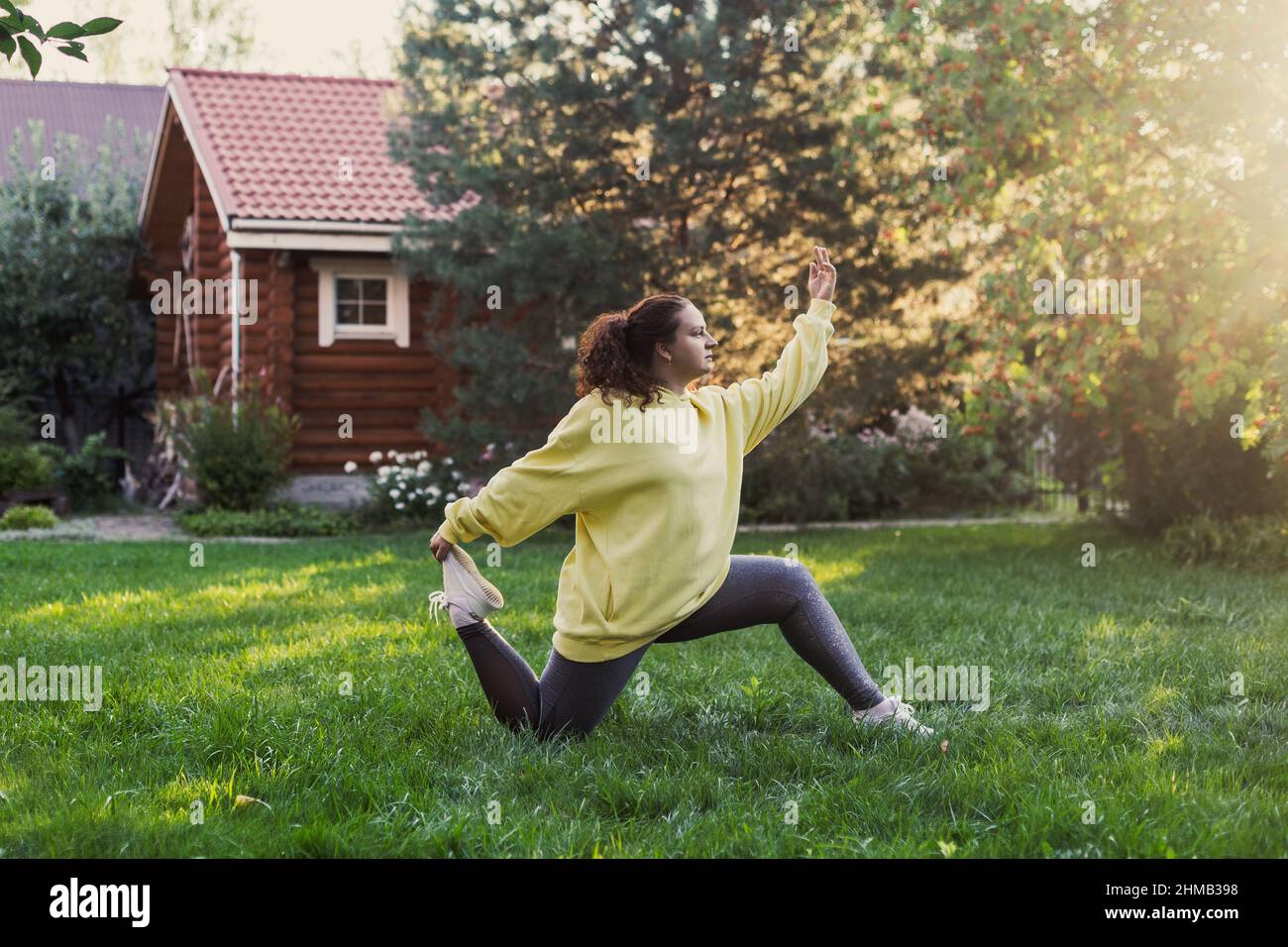 Femme caucasienne sur-pondérée dans les vêtements de sport faisant des exercices physiques sur cour sur l'herbe verte avec maison de campagne en bois et les arbres élevés dedans Banque D'Images