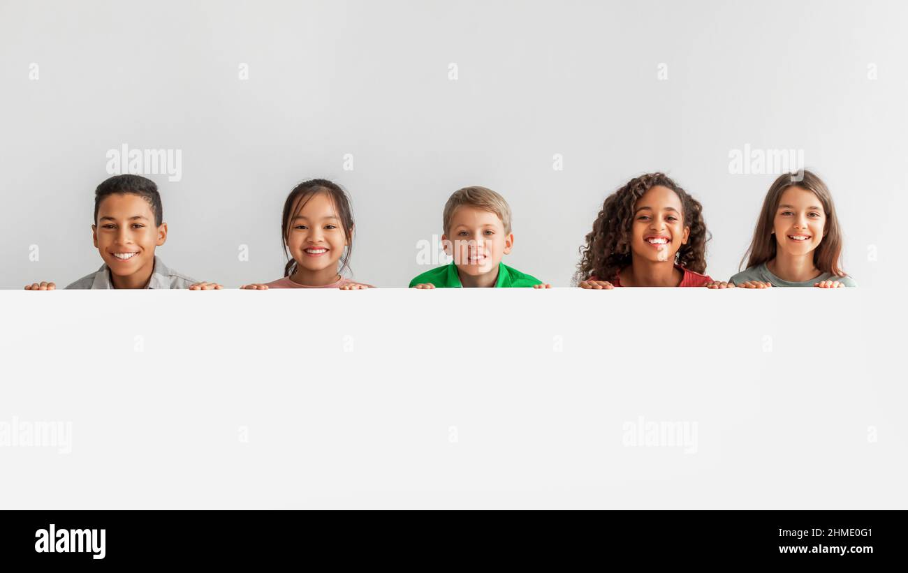 Heureux enfants multiculturels posant avec une affiche blanche vide, fond gris Banque D'Images