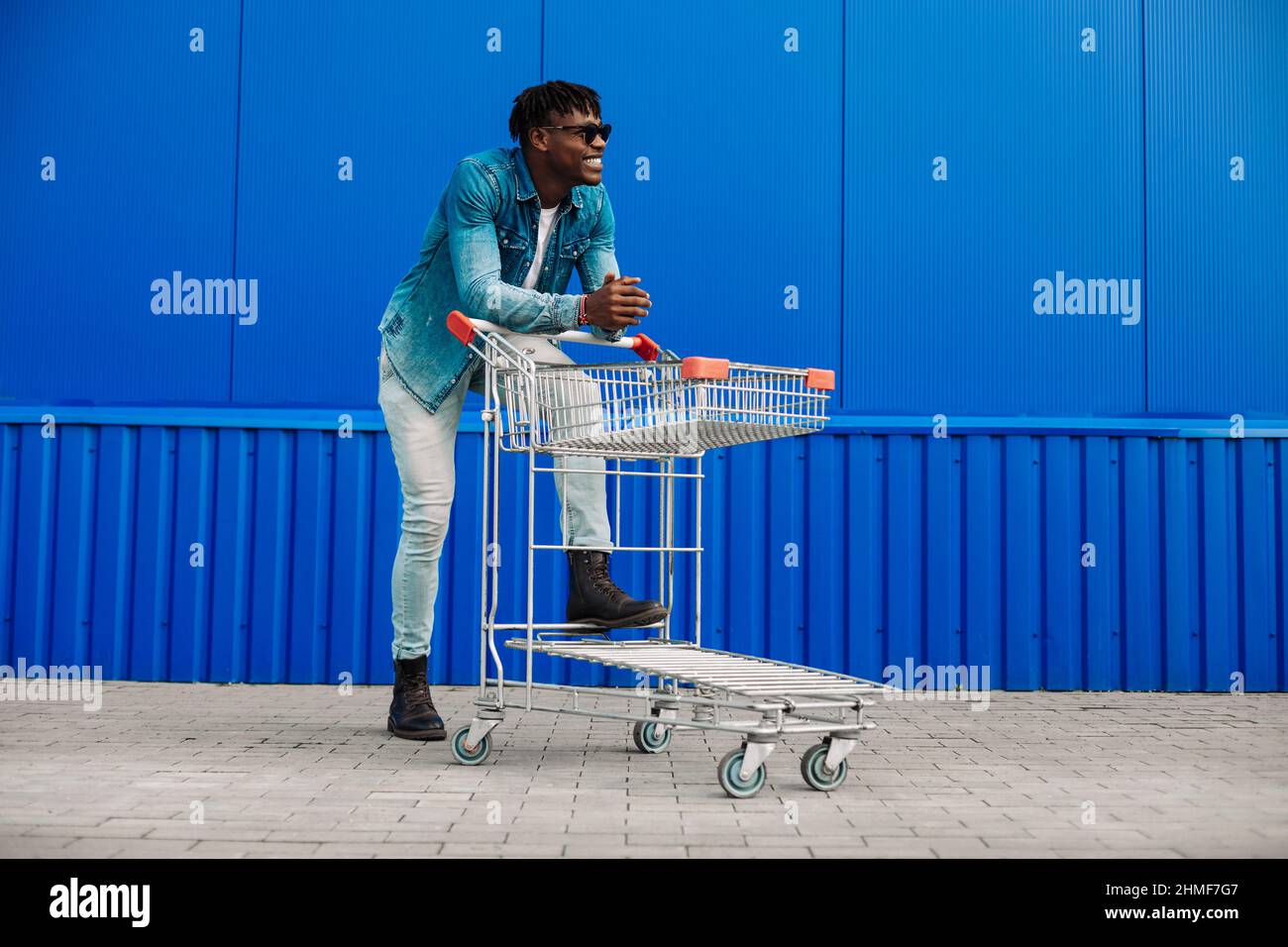 Photo du profil complet d'un homme afro-américain heureux qui fait du shopping dans des lunettes, un t-shirt, un Jean, des baskets. Un homme heureux avec un panier sur Blue wa Banque D'Images