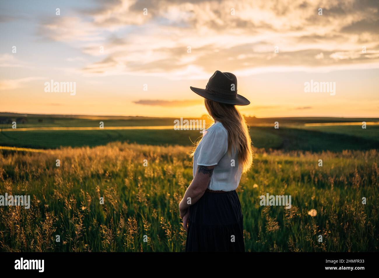 Fille en chemise blanche et chapeau profondément dans la pensée heureux avec la ferme rurale en arrière-plan au coucher du soleil Banque D'Images