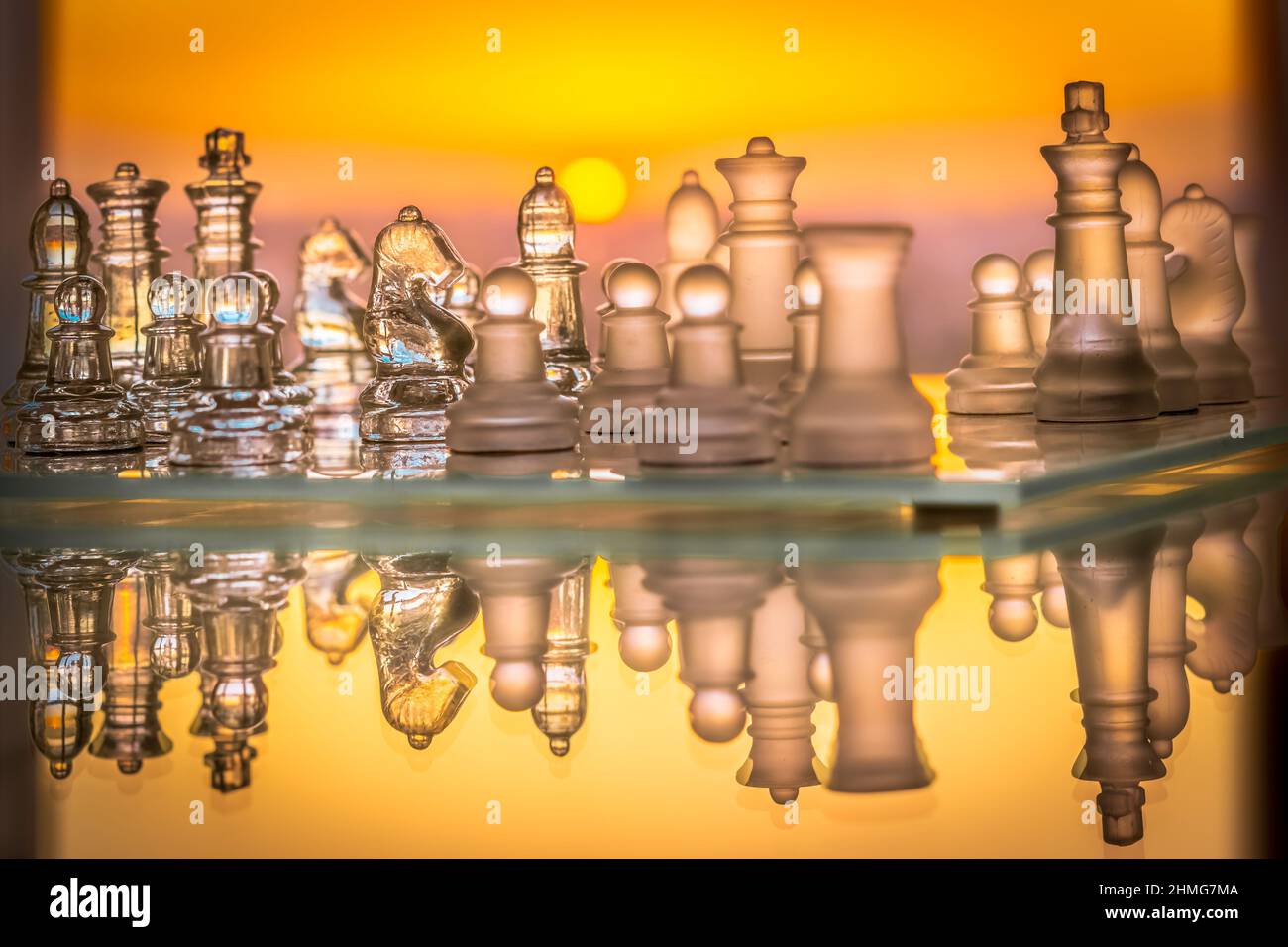 Jeu d'échecs avec pièces en verre et échiquier horizontal réfléchi avec un coucher de soleil orange saturé dans l'arrière-plan. Banque D'Images