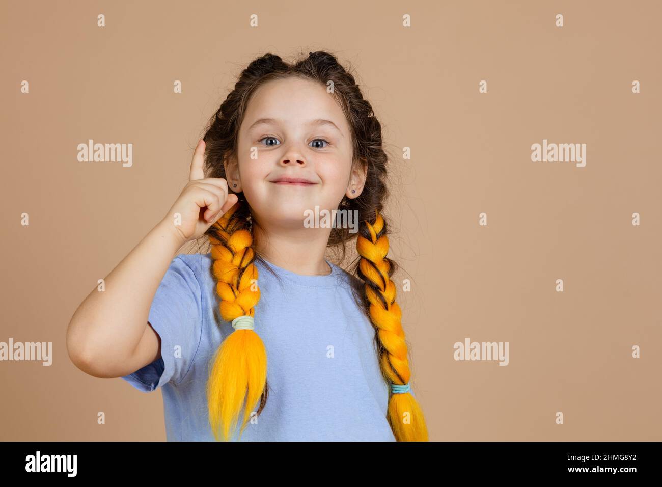 Petite fille intelligente avec des tresses kanekalon de couleur jaune montrant le doigt ayant le moment aha en raison de la grande idée apparaissant dans la tête sur fond beige Banque D'Images