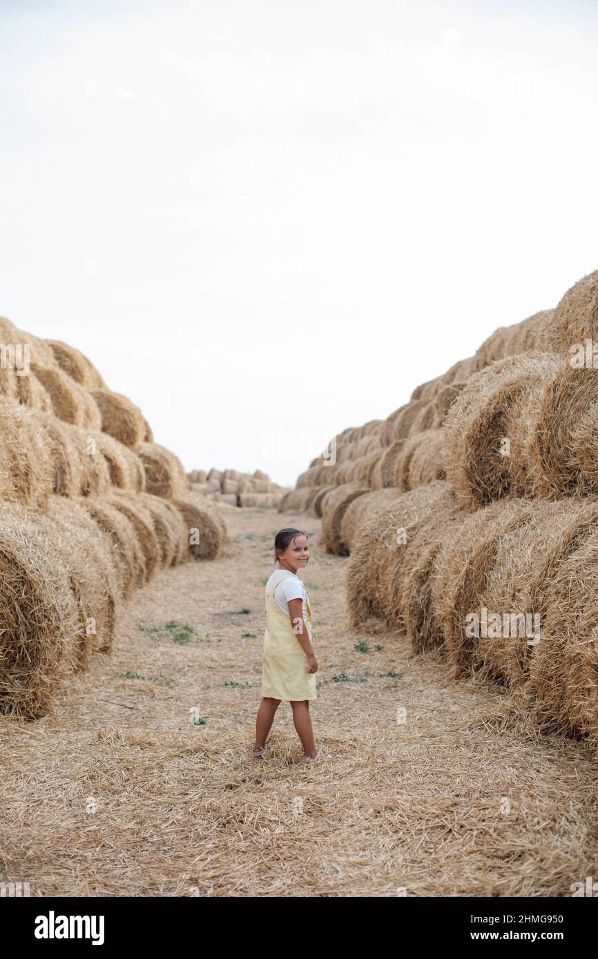 Super heureuse jeune fille souriante debout dans le foin avec dos à l'appareil photo et la tête tournée vers l'arrière regardant l'appareil photo entre les tas de foin sur le terrain dedans Banque D'Images