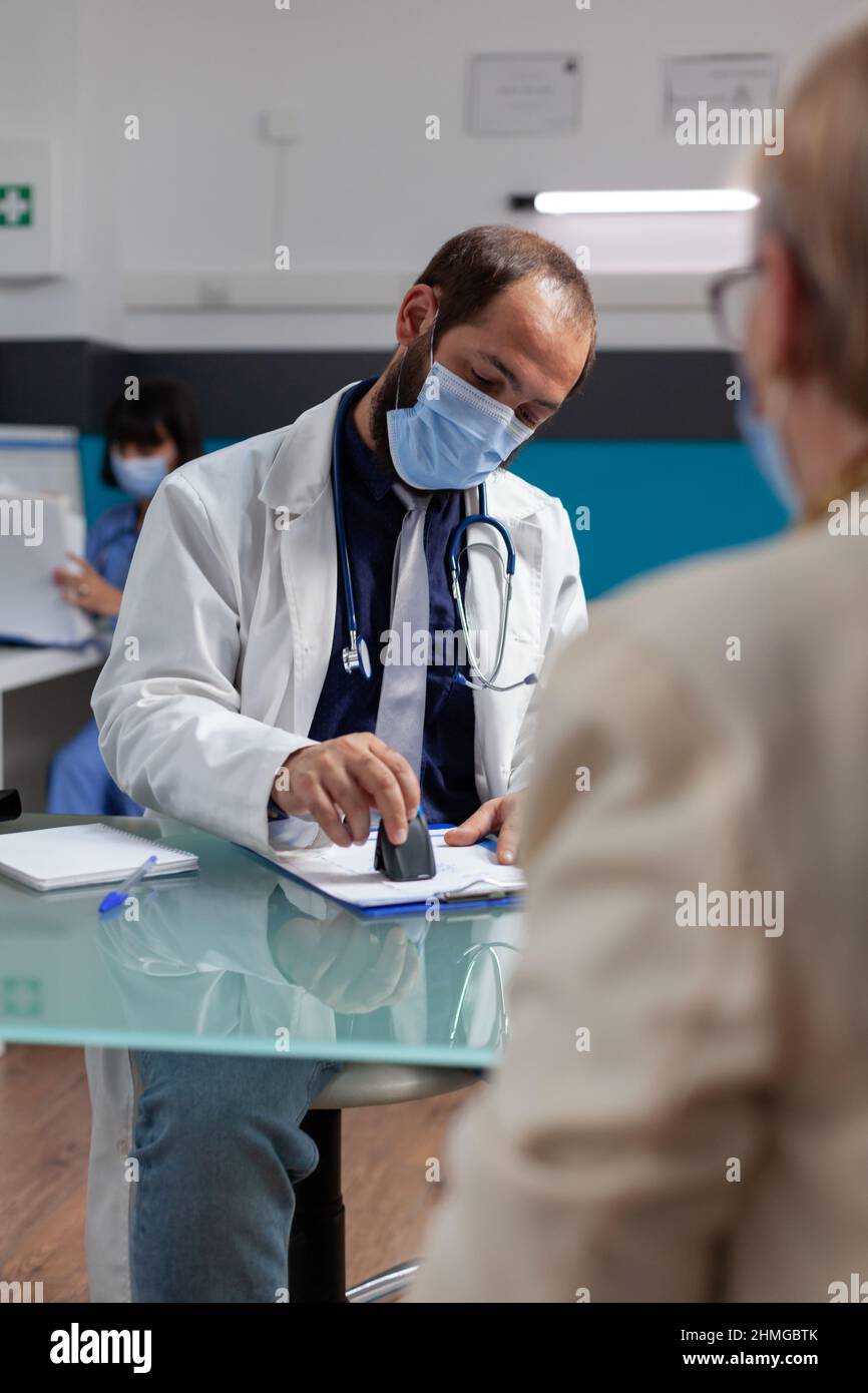 Médecin avec masque facial utilisant le timbre médical sur le papier d'ordonnance, donnant des soins de santé à la femme aînée lors de la visite de contrôle. Spécialiste en pose de sceau sur le formulaire d'examen au bureau. Banque D'Images