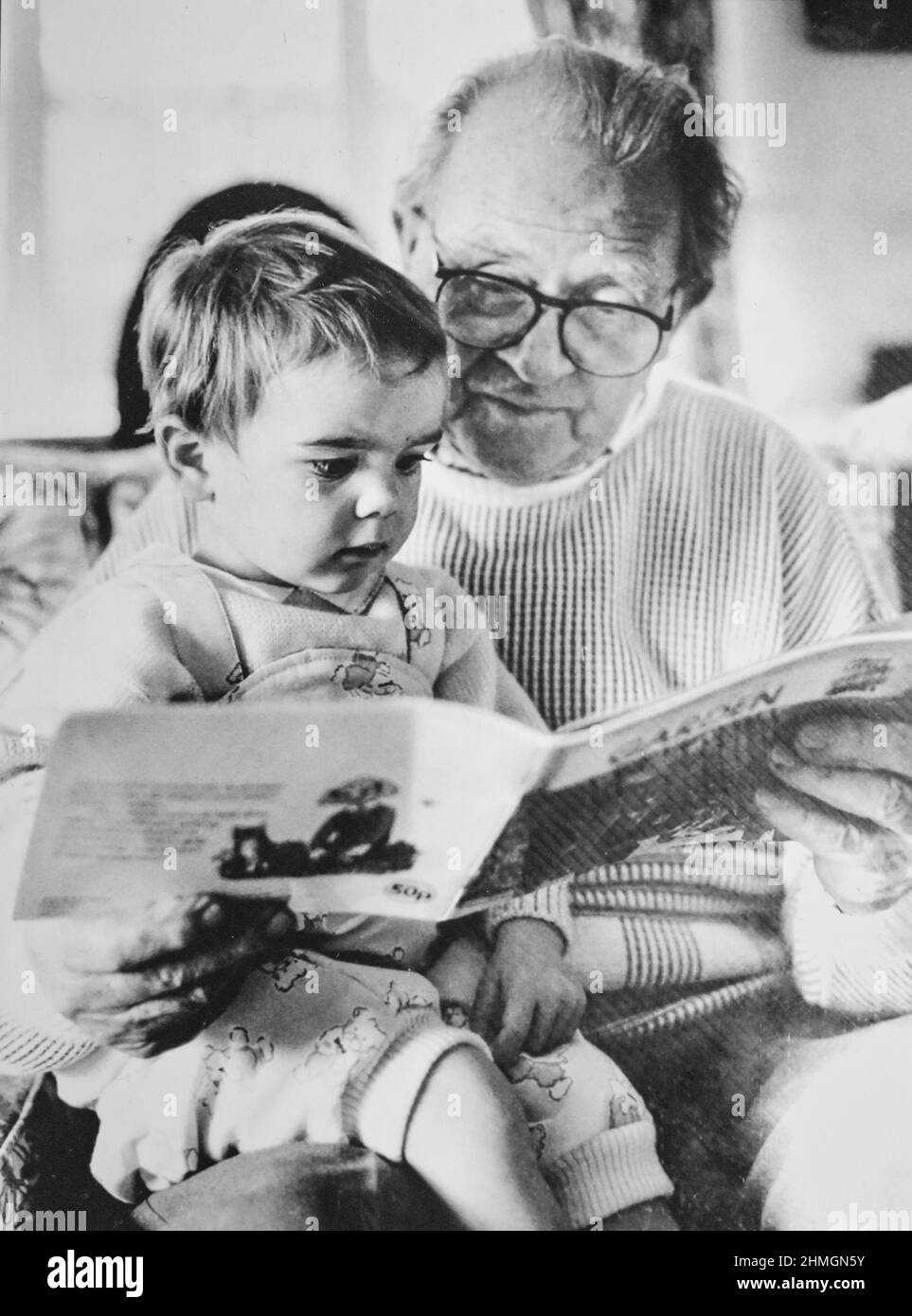 Jeune fille d'environ deux ans lisant un livre avec son grand-père 1990s Photographie prise par Simon Dack Banque D'Images