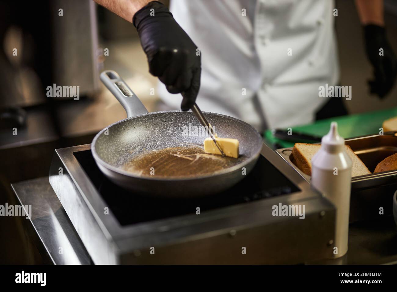 Vue en crêpe des mains du chef en gants noirs faisant fondre le beurre dans  la poêle. Homme en uniforme de chef à l'aide d'une pince à épiler pour  cuisiner avec des