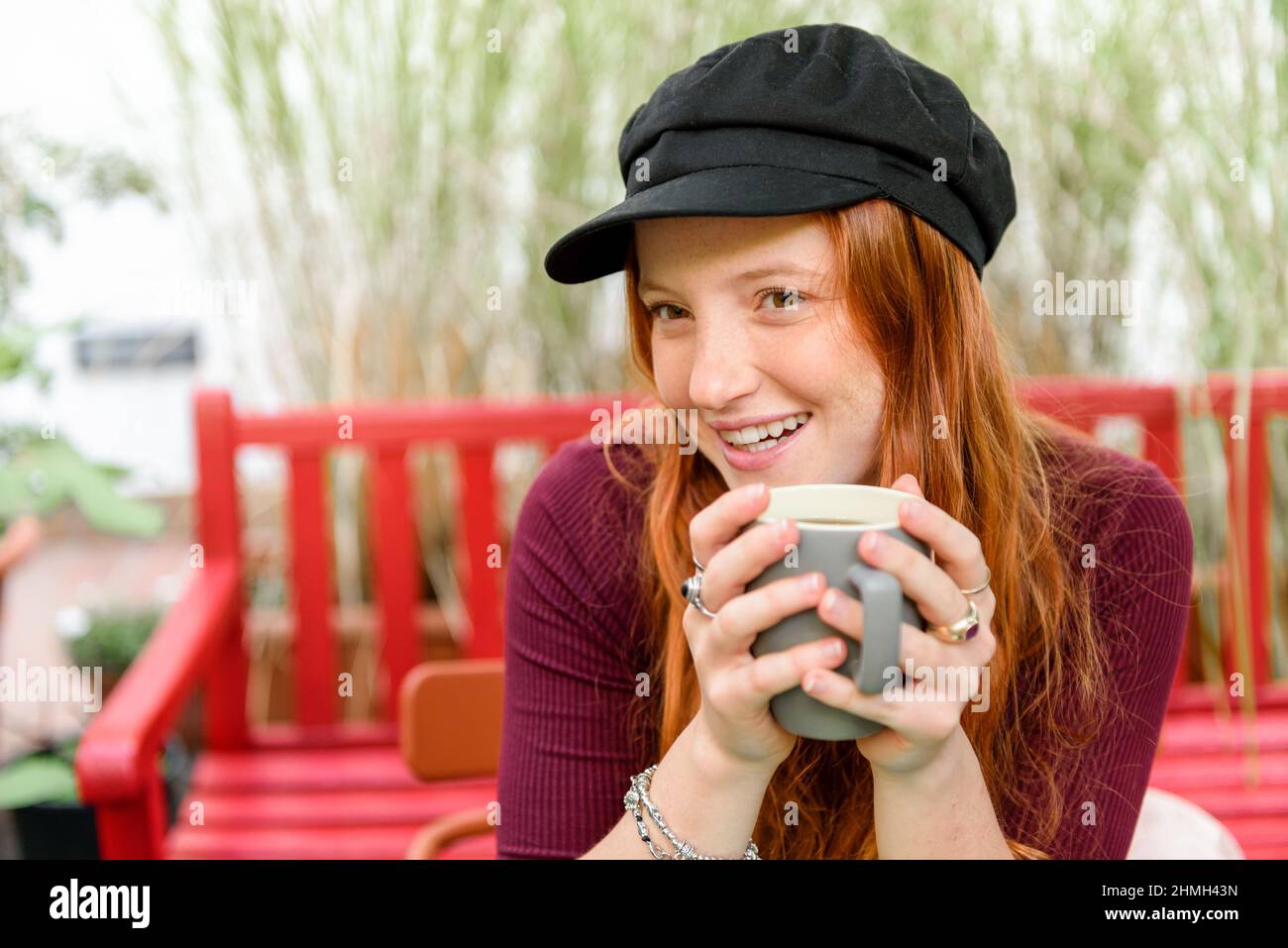 Bonne jeune femme en chapeau regardant loin en s'asseyant avec une tasse de café sur le banc dans le jardin avec des plantes vertes Banque D'Images