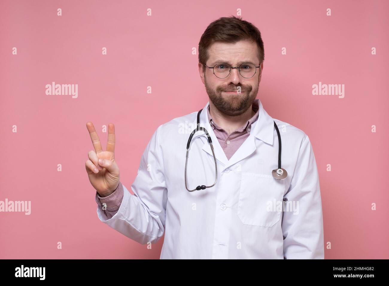 Un médecin mâle fatigué avec un stéthoscope montre un symbole de victoire. Mouvement avec deux doigts. Fond rose. Banque D'Images