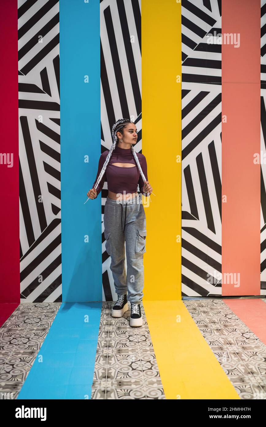 Image verticale d'une jeune femme debout embrassant ses tresses tout en regardant loin contre le mur avec des couleurs et des motifs rayés. Banque D'Images
