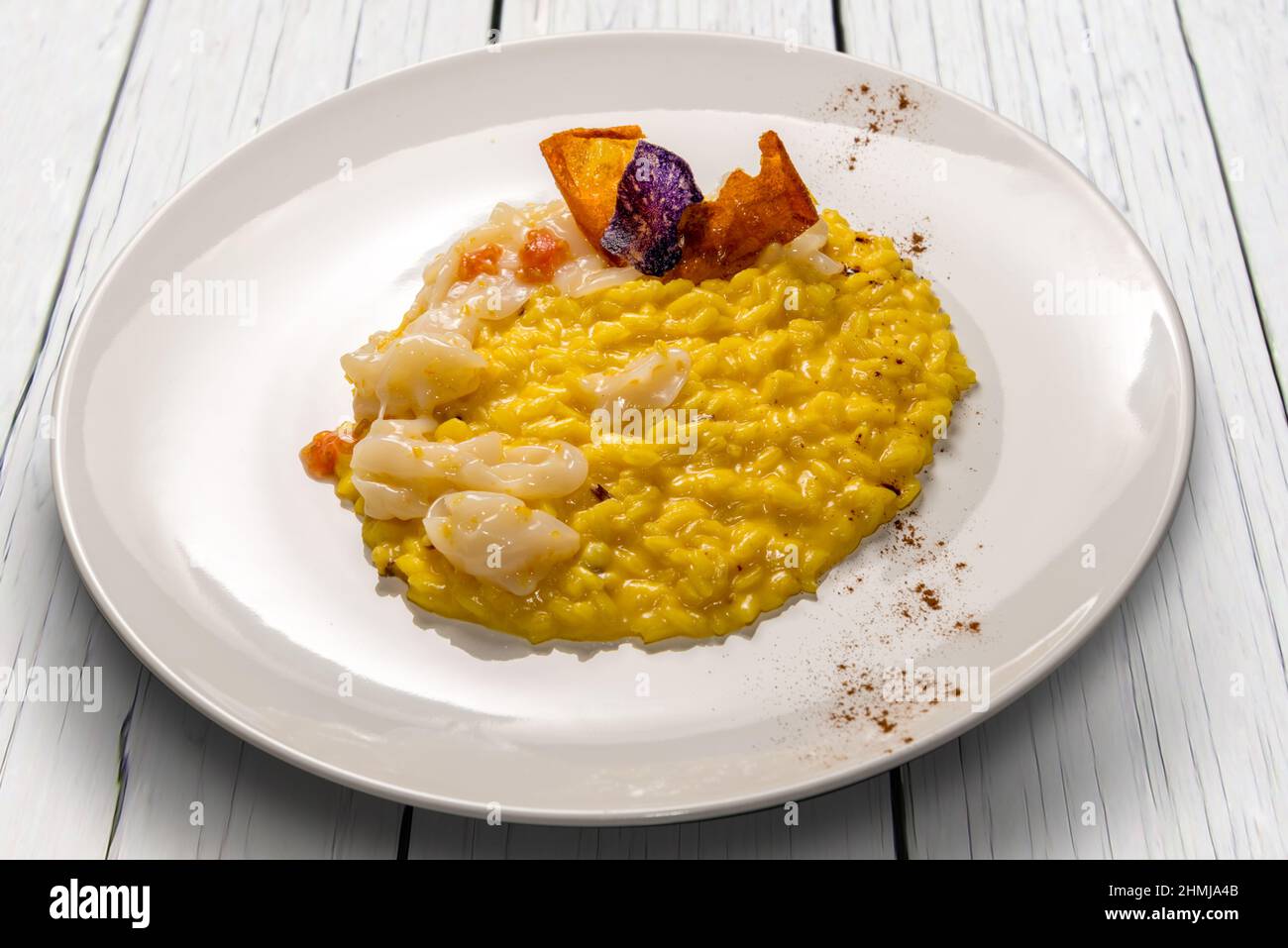 Risotto au safran jaune avec seiches et pulpe de tomate fraîche, recette gastronomique dans un plat blanc sur une table en bois blanc Banque D'Images
